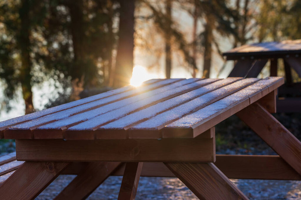 Mesa de madeira no jardim de um restaurante fechado. Há um pó de neve na mesa. O sol brilha através das árvores. - Foto, Imagem