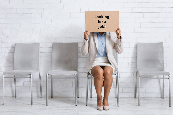 Young African American woman covering face with LOOKING FOR JOB sign while waiting for employment interview at office - Photo, Image