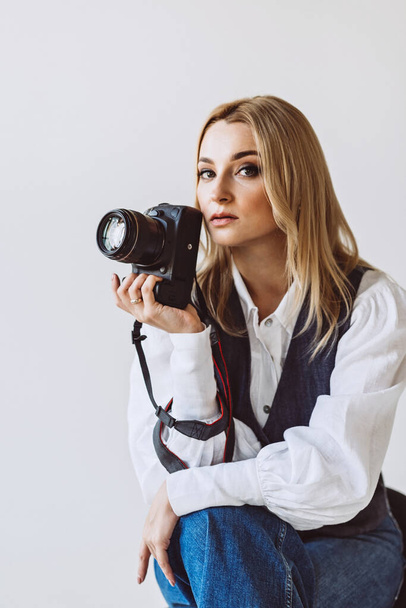 A beautiful woman photographer in a denim casual outfit and a white blouse with voluminous sleeves with a camera in her hands. Hobbies. Soft selective focus. - Foto, immagini