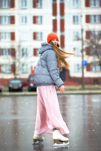 Дівчина-підліток катається на льоду в місті. Молода дівчина в довгій сукні і теплий піджак на льоду
. - Фото, зображення