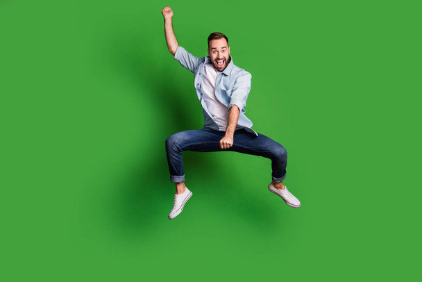 Full size photo of optimistic funky guy jump horse wear blue shirt jeans footwear isolated on green color background - Фото, изображение