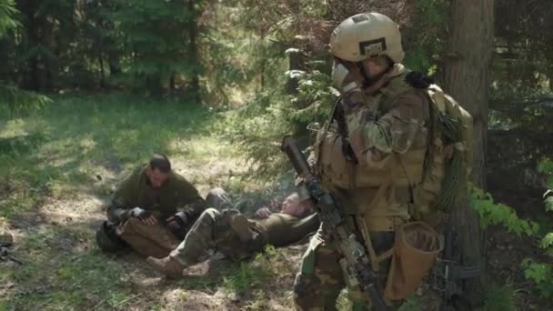 Средний выстрел воинского отряда, отдыхающего под деревьями в летнем лесу после сложной военной операции - Кадры, видео