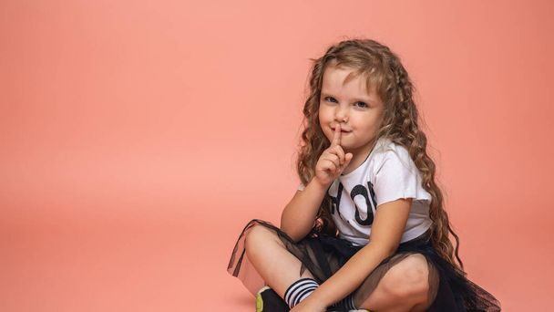 stylish little girl of 6 years old with curly hair in fashionable clothes sits on a pink background and holds a finger near her lips. Children's emotional images. - Foto, imagen