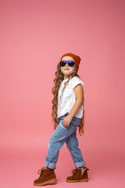 happy, stylish 6-year-old girl with curly hair in ripped jeans, a hat and sunglasses, goes full-length, isolated on a pink background. Dynamic emotional image. Copy the space bar. - Foto, Bild