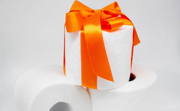 Несколько рулонов белой перфорированной туалетной бумаги. Один рулон завязан ярко-оранжевой атласной лентой в подарок. - Фото, изображение