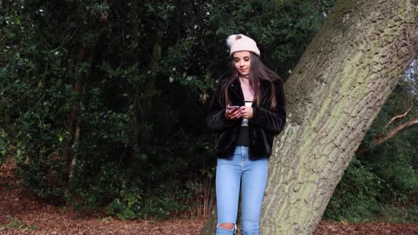 Młoda atrakcyjna kobieta w parku opierając się o drzewo w okresie zimowym, patrząc na jej telefon online dating przesuwając w lewo iw prawo na aplikacji randkowej, lubiąc niektóre profile i nie lubiąc niektóre - Materiał filmowy, wideo