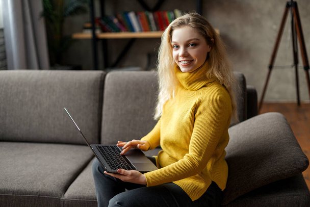 Beautiful young woman working from home on a laptop sitting on the sofa in the living room - Φωτογραφία, εικόνα