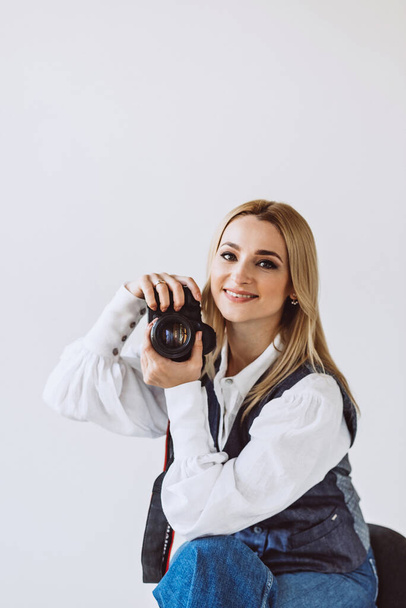 A beautiful woman photographer in a denim casual outfit and a white blouse with voluminous sleeves with a camera in her hands. Hobbies. Soft selective focus. - Фото, зображення