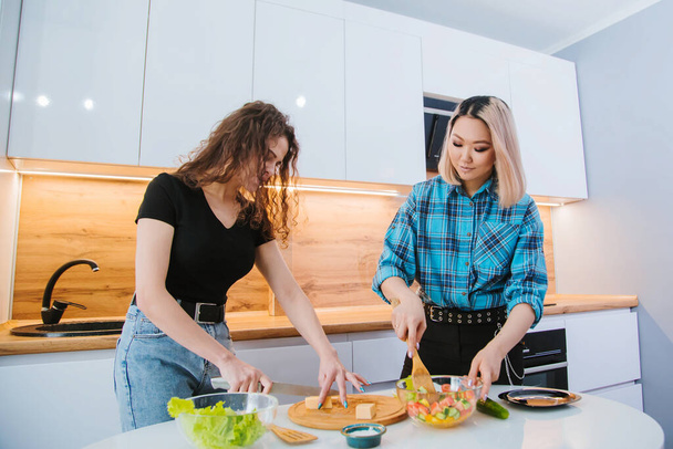 2人の女の子の友人は、軽いキッチンを背景に食事のサラダを準備しています。健康的な食事. - 写真・画像
