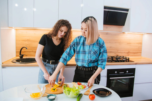 2人の女の子の友人は、軽いキッチンを背景に食事のサラダを準備しています。健康的な食事. - 写真・画像
