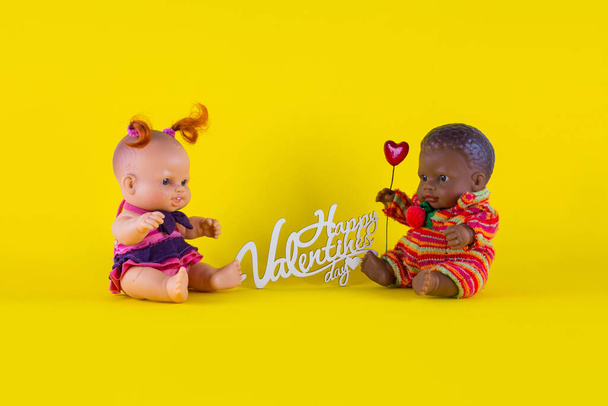 Valentine 's Day card Dolls - una chica blanca con rizos rojos y un chico negro con un corazón están sentados en un fondo amarillo uno enfrente del otro - Foto, imagen