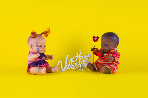 Valentine 's Day card Dolls - una chica blanca con rizos rojos con una ramita de lavanda y un chico negro con un corazón están sentados en un fondo amarillo uno enfrente del otro - Foto, Imagen