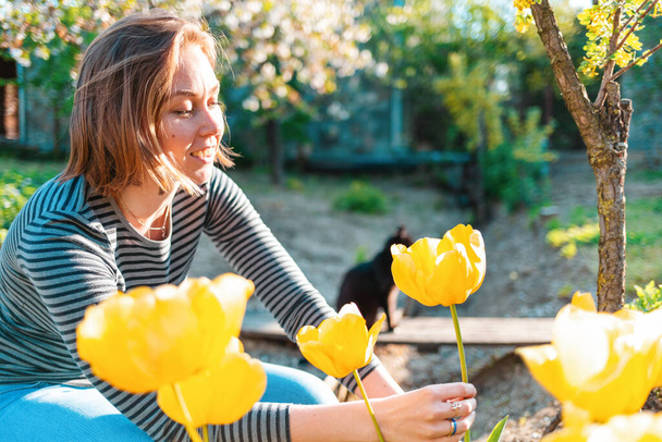 Femme souriante blonde caucasienne assise et coupant des tulipes jaunes dans le jardin. Au fond, un jardin ensoleillé et un chat. - Photo, image