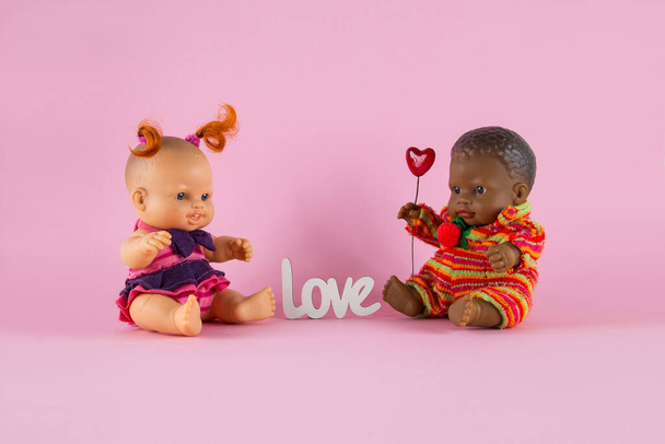 Valentin napi kártya Dolls - egy fehér lány piros fürtök és egy fekete fiú egy szív ül a rózsaszín háttér egymással szemben - Fotó, kép