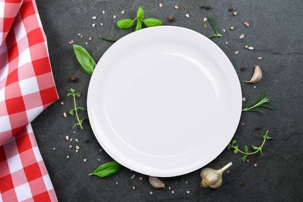 Класичний італійський кулінарний шаблон - вид білої пустої керамічної тарілки на старовинному кам'яному кухонному стільнику з червоною пробною скатертиною та вибором трав і спецій.. - Фото, зображення