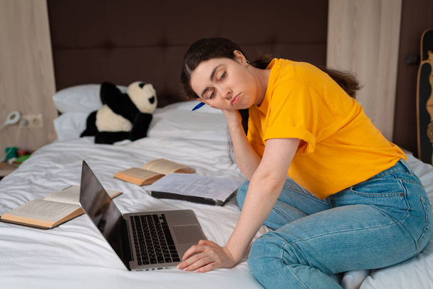 Concept of online courses and online education. Portrait of a young Caucasian woman is sitting with a bored look on the bed, and a laptop and books are lying next to her. - Φωτογραφία, εικόνα