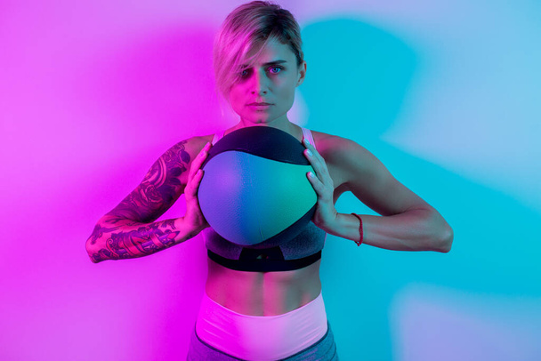 Fit young blonde woman exercise with medicine ball in gym in neon lights. - Zdjęcie, obraz