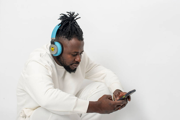 Waist up portrait of serious dark skinned African male with pigtails and in blue headphones wearing white tracksuit looking at phone against white baskground in studio - Photo, Image