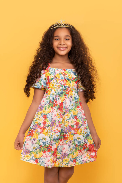 Portrait demi-longueur d'une jeune fille à la peau foncée avec de longs cheveux bouclés portant une robe colorée sur fond jaune - Photo, image