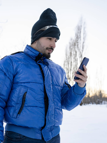 όμορφος άνδρας κοιτάζει κάμερα, χρησιμοποιώντας το κινητό τηλέφωνο σε μια ηλιόλουστη μέρα, χιόνι φόντο - Φωτογραφία, εικόνα