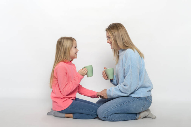 Aufnahme von gut gelaunten Mutter und Tochter mit Tassen Tee, die auf dem Boden sitzen und sich vor grauem Hintergrund freundlich unterhalten. - Foto, Bild