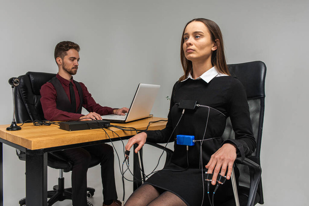 Concentrated, thoughtful woman is in a bright room, testing on a computer polygraph. Young man sitting at the table and looking at the polygraph screen and polygraph monitoring. The concept of truth, polygraph - Foto, Imagem