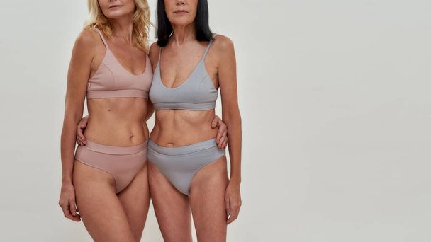 Cropped shot of two caucasian women in underwear looking at camera while posing together over light background. Warmth in relation, mature beauty concept - Fotó, kép