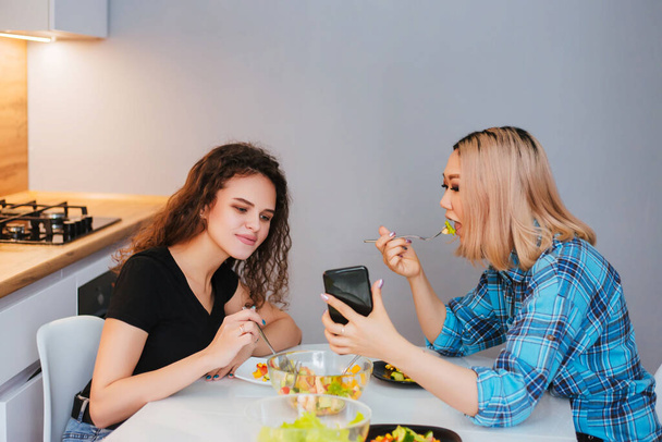 Két lány ül a konyhaasztalnál, diétás salátát esznek és videót néznek egy okostelefonon. A barátnők beszélgetnek étkezés közben. Egészséges táplálkozás - Fotó, kép