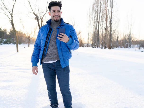 handsome smiling man, wearing coat in the snow on a sunny day at sunset, with his smartphone in hand - Foto, Bild