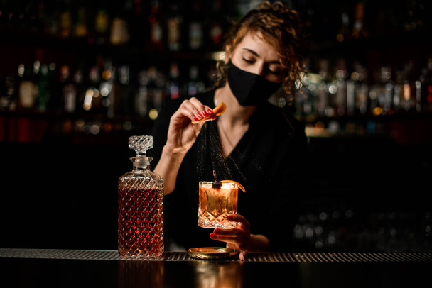 view on woman bartender in mask who holds glass of cocktail and sprinkles on it - Foto, Imagen