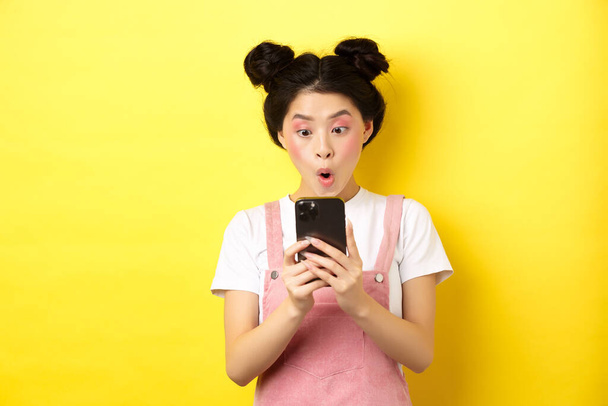 Азиатская девочка-подросток смотрит взволнованно на экран смартфона, читает новости по телефону, стоя на желтом фоне - Фото, изображение