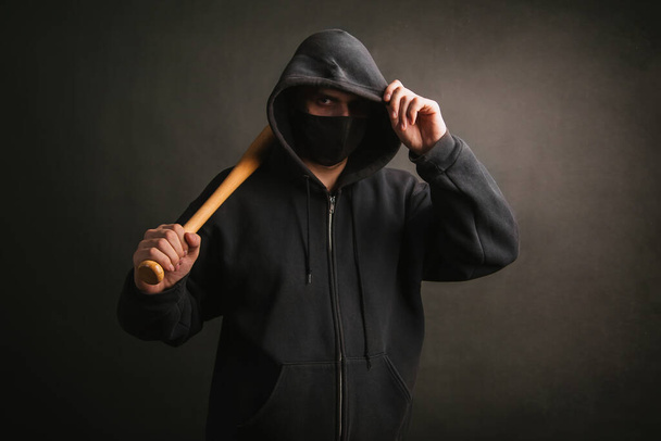 Bandit in a black sweatshirt and a medical mask. Dangerous man with a baseball bat ready to fight. The guy goes to protest - Фото, изображение
