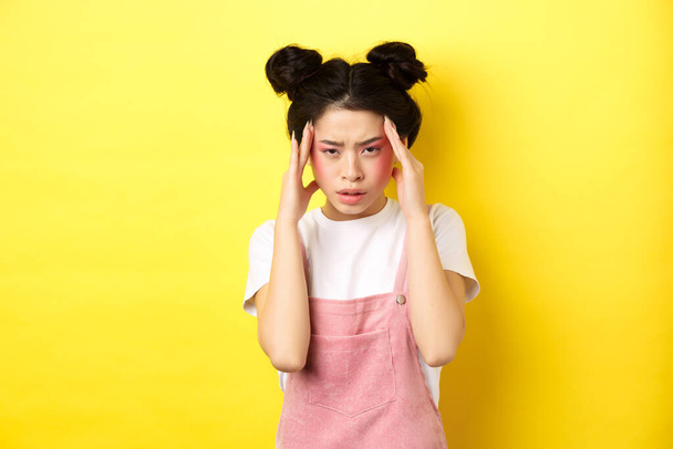 Image of stylish japanese woman with bright makeup, having headache, touching head and looking tired, have painful migraine, yellow background - Foto, afbeelding