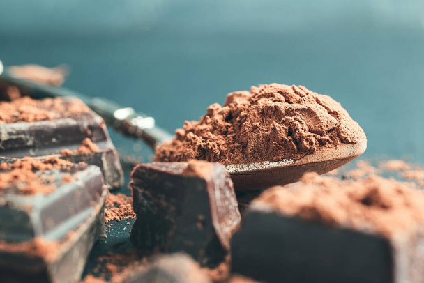 Коричневый какао порошок в ложке и рубленые кубики шоколада на темном фоне с копировальным местом. - Фото, изображение
