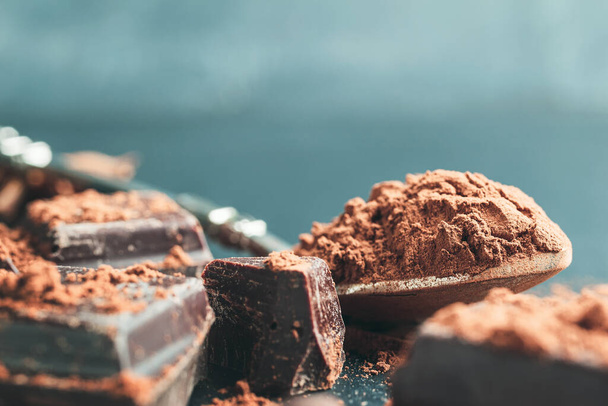 Polvo de cacao marrón en la cuchara y cubos de chocolate picados sobre fondo oscuro con espacio para copiar. - Foto, imagen