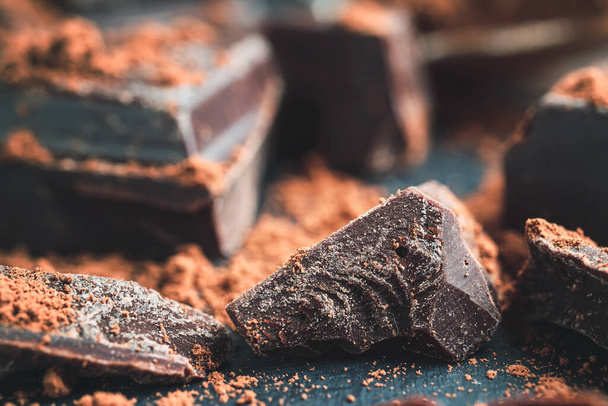 Trozo de chocolate, cubos de chocolate picados con cacao en polvo sobre fondo oscuro, vista de cerca. - Foto, imagen