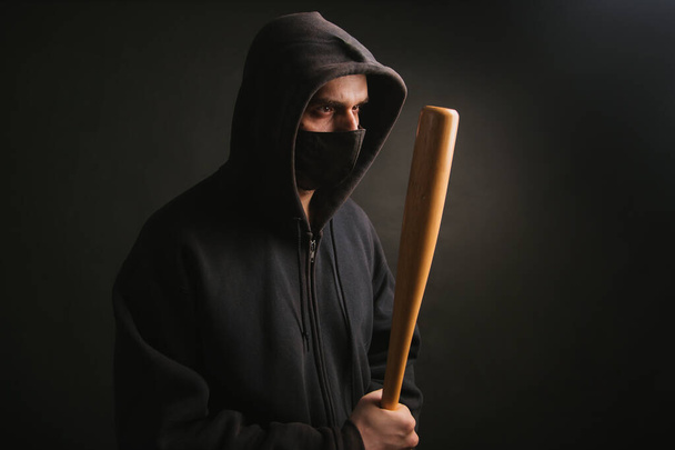 Bandido con una sudadera negra y una máscara médica. Un hombre peligroso con un bate de béisbol listo para luchar. El tipo va a protestar. - Foto, Imagen