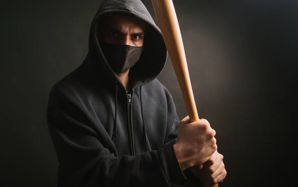 Bandit in schwarzem Sweatshirt und medizinischer Maske. Gefährlicher Mann mit kampfbereitem Baseballschläger. Der Typ geht zum Protest - Foto, Bild