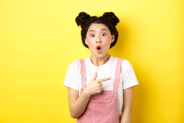 Excited stylish asian woman with glamour makeup, pointing finger right and gasping fascinated, say wow amazed, yellow background - Photo, image