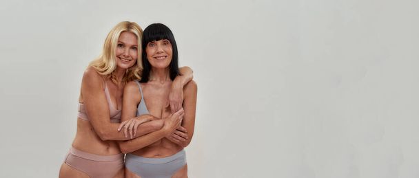Portrait of two women in underwear smiling at camera, embracing each other while posing together over light background. Warmth in relation, mature beauty concept - Foto, afbeelding