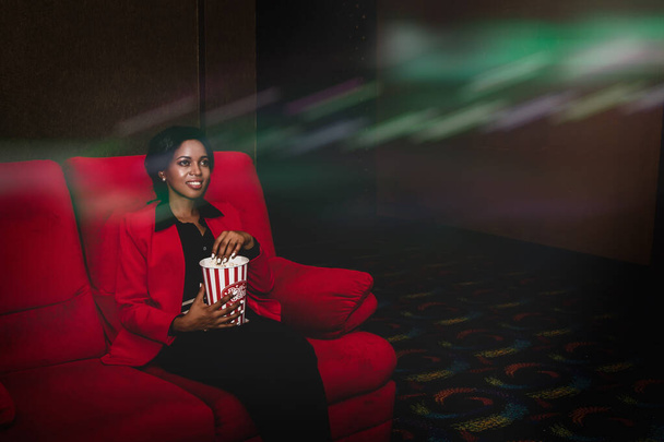 Красивая афро-американка сидит и смотрит кино одна на красном диване и забирает немного консервированного попкорна, чтобы поесть, как прекрасное время, чтобы посмотреть ее развлечения. - Фото, изображение