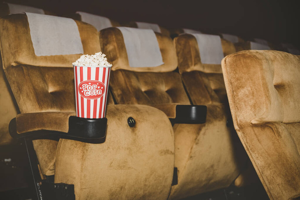 Popcorn συσκευάζεται σε ένα κόκκινο χάρτινο κύπελλο τοποθετείται σε μια καρέκλα σε έναν κινηματογράφο τρώγεται για πρόσθετη απόλαυση. - Φωτογραφία, εικόνα