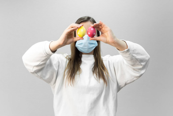 Woman with Easter eggs on eyes and medical mask on face. Easter during the coronavirus pandemic. Safety at Easter 2021 in quarantine - Foto, immagini