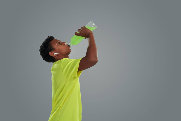 More energy. Side view of a teenage african boy wearing sports clothes drinking energy drink while standing against grey background - Foto, immagini