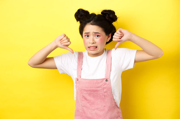 Disgusted asian girl showing thumbs down and cringe from bad product, standing in summer clothes against pink background - Photo, Image