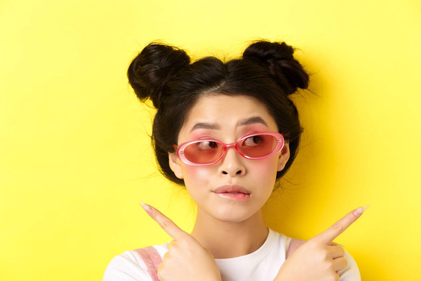 Καλοκαιρινή μόδα. Pensive κορίτσι της Ασίας κάνει την επιλογή, δείχνοντας προς τα πλάγια και κοιτάζοντας στοχαστική, να αποφασίσει τι επιλογή, στέκεται με γυαλιά ηλίου σε κίτρινο φόντο - Φωτογραφία, εικόνα
