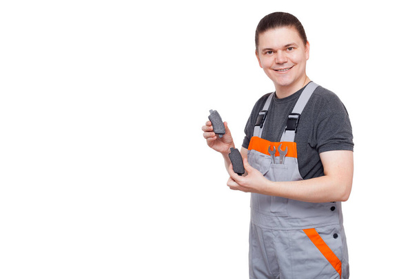 male employee in gray uniform of car mechanic worker with spanner wrench in his pocket holds brake pads in his hands looks into camera and smiles isolated on white background man with car spare parts. - 写真・画像