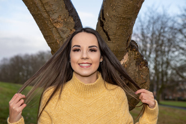 A young attractive woman with long brown hair in the winter time in a wooden park area wearing a yellow jumper looking by a tree in the forest - Photo, Image