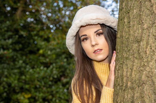Una joven atractiva mujer con el pelo largo y castaño en invierno en un parque de madera con un jersey amarillo mirando desde detrás de un árbol. - Foto, Imagen
