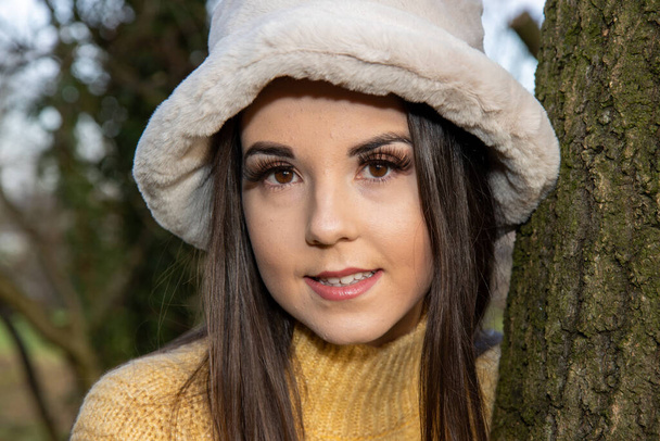 Egy fiatal, vonzó nő hosszú barna hajjal télen egy fapark területén sárga pulóvert visel, ami egy fa mögül néz ki.. - Fotó, kép
